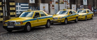 Three yellow taxis with blue stripes are parked in a row on a cobblestone street. They are all Mercedes-Benz models and have the word 'TAXI' displayed on their roofs. The background features a wall with various painted murals and patterns.