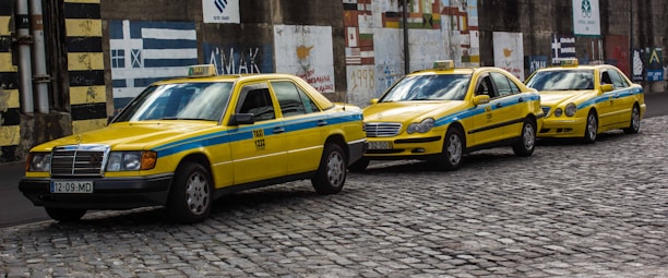 Three yellow taxis with blue stripes are parked in a row on a cobblestone street. They are all Mercedes-Benz models and have the word 'TAXI' displayed on their roofs. The background features a wall with various painted murals and patterns.