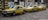 Three yellow taxis with blue stripes are parked in a row on a cobblestone street. They are all Mercedes-Benz models and have the word 'TAXI' displayed on their roofs. The background features a wall with various painted murals and patterns.