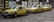Three yellow taxis with blue stripes are parked in a row on a cobblestone street. They are all Mercedes-Benz models and have the word 'TAXI' displayed on their roofs. The background features a wall with various painted murals and patterns.