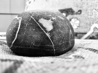 A smooth, round stone with visible white veins rests on a textured surface. The background shows a blurred fabric or textile pattern with geometric shapes. The image is in black and white, highlighting contrasts and textures.