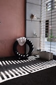 A black tire leaned against a wall with a piece of cloth draped over it. There is a metal tap fixture on a tiled wall, along with a cinder block on the ground. Sunlight is casting shadows through a metal grate onto the concrete floor.