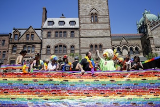A group of people with colorful costumes and wigs stand behind a vibrant rainbow banner in front of historic stone buildings with gothic architecture. The scene is bathed in bright sunlight.