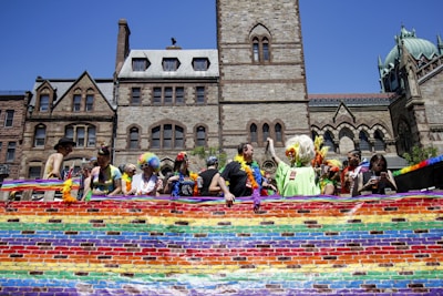 A group of people with colorful costumes and wigs stand behind a vibrant rainbow banner in front of historic stone buildings with gothic architecture. The scene is bathed in bright sunlight.