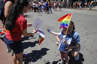 A young child holding a rainbow flag is interacting with an adult, possibly receiving or giving items, during a street event. In the background, a larger crowd can be seen, some holding banners and wearing festive attire. The ground is scattered with colorful confetti, adding vibrancy to the scene.