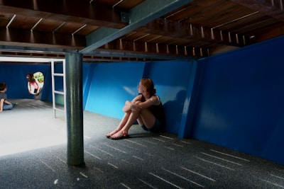 A young person sits alone under a structure with a blue wall and carpeted floor, looking contemplative. Light streams in from an opening on the left, where two other children, one sitting and one entering through a round hole, are engaged in the background.