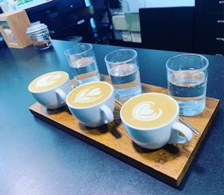 A wooden tray holds three cups of latte art next to three glasses of water, arranged neatly in a row. The tabletop is dark, contrasting with the white cups and clear glasses. Background elements include jars and kitchen items slightly blurred.