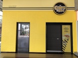 A yellow building facade features a closed black shutter door and a glass door. Above the shutter is a sign that reads 'CHIPS & DIPS' with a logo. Next to the black shutter, a poster shows an image of fries and the text 'GREAT FRIES! GREAT TIME!'. The building has a textured brick pattern on the wall and includes some Arabic text.