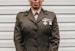 A person is wearing a military uniform with several medals attached to the chest. The background consists of light-colored horizontal panels.
