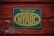 A weathered sign attached to a rustic, red-painted wooden surface. The sign is rectangular with a dark green background and a faded yellow oval around the letters 'NYABC'. It contains text that reads 'Technician New York Artificial Breeders' Cooperative, Inc.' The wooden texture shows signs of peeling paint and age.