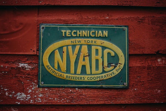 A weathered sign attached to a rustic, red-painted wooden surface. The sign is rectangular with a dark green background and a faded yellow oval around the letters 'NYABC'. It contains text that reads 'Technician New York Artificial Breeders' Cooperative, Inc.' The wooden texture shows signs of peeling paint and age.