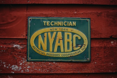 A weathered sign attached to a rustic, red-painted wooden surface. The sign is rectangular with a dark green background and a faded yellow oval around the letters 'NYABC'. It contains text that reads 'Technician New York Artificial Breeders' Cooperative, Inc.' The wooden texture shows signs of peeling paint and age.