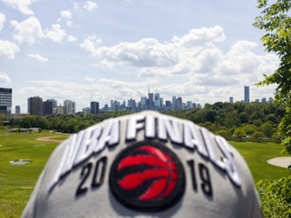 A city skyline is visible in the distance under a partially cloudy sky. In the foreground, a cap with the text 'NBA FINALS 2019' and a red basketball logo is prominently displayed. The scene includes a mix of high-rise buildings, green parkland, and trees.
