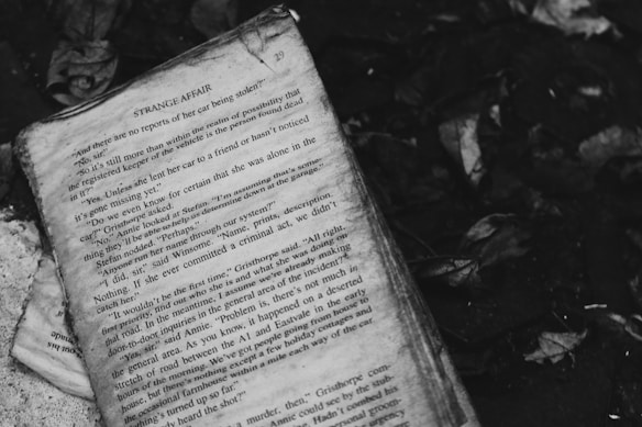 An old, weathered book page rests against a dark background scattered with dried leaves. The text on the page is partially legible, suggesting a mystery or crime story.