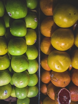 A close-up display of neatly stacked fresh green apples and ripe oranges. The apples are on the left side, while the oranges are on the right. The fruits have small labels attached to them, indicating they are store-bought.