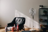 Coke soda bottles near vacant black armchair beside white pedestal fan inside white room