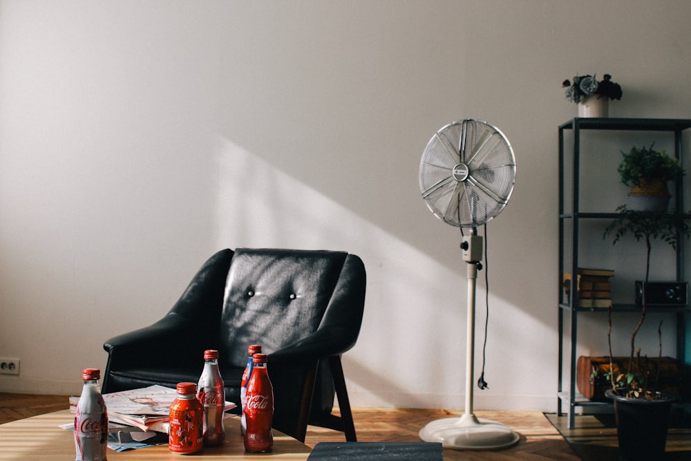 Coke soda bottles near vacant black armchair beside white pedestal fan inside white room