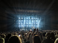 A concert scene with a large audience, where numerous people hold up phones to capture the performance. Bright stage lights create starburst patterns, illuminating the stage and crowd, while a band performs.