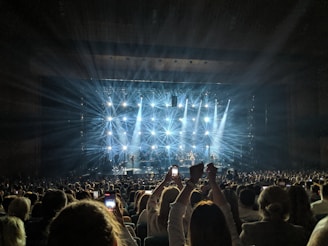 A concert scene with a large audience, where numerous people hold up phones to capture the performance. Bright stage lights create starburst patterns, illuminating the stage and crowd, while a band performs.