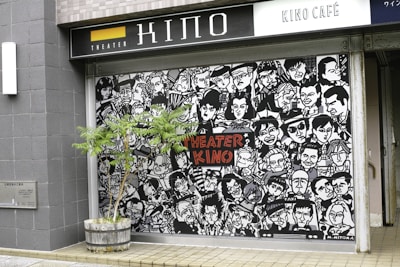 A mural featuring numerous stylized black-and-white portraits with expressive faces covers the front of a building. The words 'Theater Kino' are prominently displayed in red letters in the center. A small tree in a wooden planter is positioned in front of the mural. The scene is set on a tiled pavement.