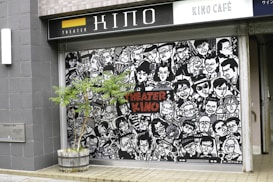 A mural featuring numerous stylized black-and-white portraits with expressive faces covers the front of a building. The words 'Theater Kino' are prominently displayed in red letters in the center. A small tree in a wooden planter is positioned in front of the mural. The scene is set on a tiled pavement.
