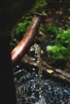 A rusted metal pipe is positioned over a surface with clear water flowing freely from it. In the background, blurred greenery provides a natural setting.