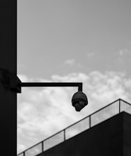 A surveillance camera is mounted on a wall with a stark, industrial design. The background consists of a cloudy sky and a large building edge, giving off a minimalist, urban feel.