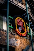 A neon sign hangs against the backdrop of a brick building, featuring bright colors in the shape of a stylized woman with a cocktail glass. The surrounding architecture includes metal frameworks and staircases, adding an urban aesthetic.