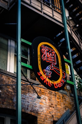 A neon sign hangs against the backdrop of a brick building, featuring bright colors in the shape of a stylized woman with a cocktail glass. The surrounding architecture includes metal frameworks and staircases, adding an urban aesthetic.
