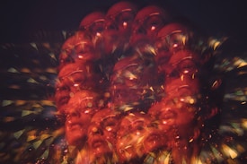 An abstract, kaleidoscopic design featuring multiple overlapping faces of a person, with glasses visible. The image is suffused with warm hues of red and orange, interspersed with glittering light effects and geometric patterns, creating a vibrant, surreal appearance.