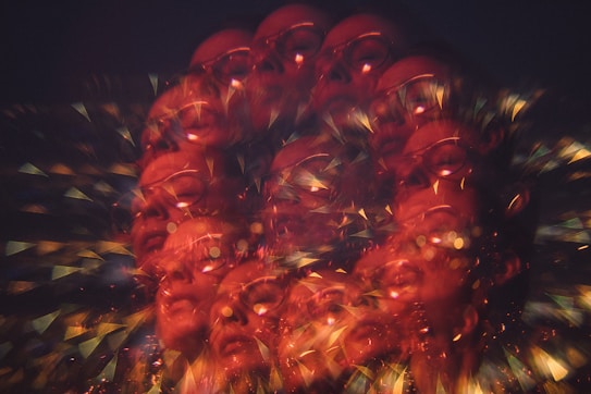 An abstract, kaleidoscopic design featuring multiple overlapping faces of a person, with glasses visible. The image is suffused with warm hues of red and orange, interspersed with glittering light effects and geometric patterns, creating a vibrant, surreal appearance.
