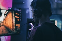 A person wearing headphones is engaged in gaming on a computer, with a large screen displaying a video game scene, showing a wooden building and a path.