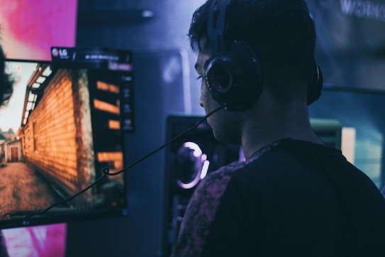 A person wearing headphones is engaged in gaming on a computer, with a large screen displaying a video game scene, showing a wooden building and a path.