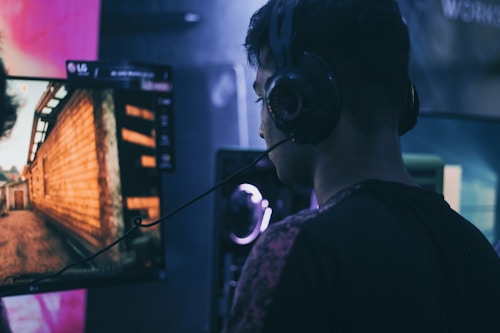 A person wearing headphones is engaged in gaming on a computer, with a large screen displaying a video game scene, showing a wooden building and a path.