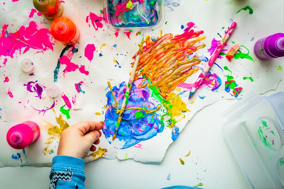 Close-up of a child's left hand with painting materials scattered across a white table and canvas.