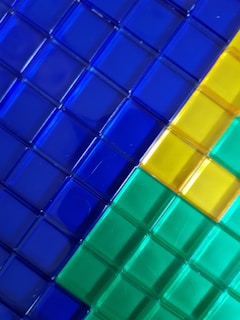 A close-up view of several transparent, glossy square tiles arranged in a diagonal pattern. The tiles are in vibrant colors including blue, yellow, green, and cyan, with reflections visible on the surface.