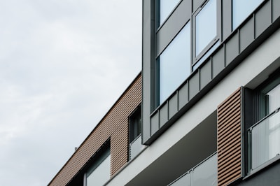 A modern building facade with large glass windows and stylish horizontal wooden slats. The architecture features clean lines and a minimalist design, highlighted by a combination of gray and neutral tones.