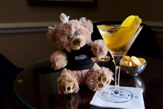 A plush teddy bear wearing a black shirt is seated at a table next to a martini glass with a lemon twist garnish. There is a bowl of crackers and a bowl of snacks in the background, along with a napkin bearing the logo 'DUKES LONDON'. The setting suggests a cozy, upscale environment.