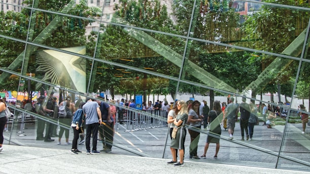 Reflections on a glass building with trees and people visible through the panes, creating a kaleidoscopic effect. The scene features groups of people walking and interacting, with lines and queues formed outside. The urban environment is bustling, with greenery from trees mirrored in the glass facade.