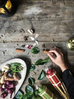 A rustic wooden table is adorned with various culinary ingredients and decorative elements. A hand reaches in to hold a cinnamon stick, surrounded by spices like star anise, garlic, and peppercorns. A plate displays a selection of fresh ingredients including green chilies, shallots, and ginger. Beside it, tropical leaves and traditional decorative items, possibly torches or lanterns, enhance the aesthetic. The setting suggests a theme of natural and traditional cooking or celebration.