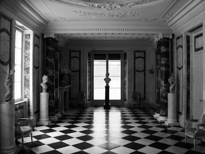 An elegant, spacious room with a black and white checkered marble floor, ornate plaster ceiling details, and marble columns. Several classical sculptures are placed around the room, with large windows letting in natural light. The decor is opulent and classical.