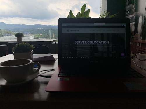 A dimly lit indoor setting featuring a laptop with a webpage titled 'Server Colocation' on the screen. In the foreground, a cup of coffee sits on a wooden table beside a potted plant. Through the window in the background, green hills and a cloudy sky are visible, providing a contrast to the darker interior.