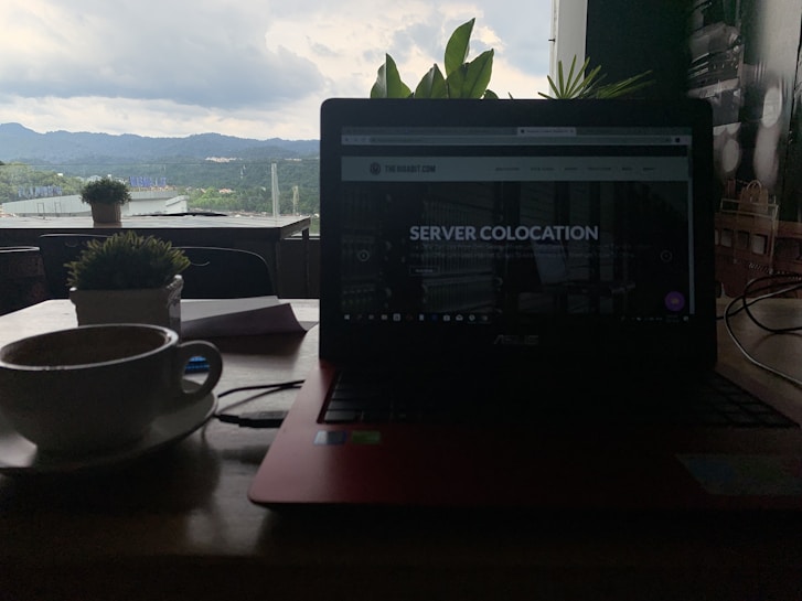 A dimly lit indoor setting featuring a laptop with a webpage titled 'Server Colocation' on the screen. In the foreground, a cup of coffee sits on a wooden table beside a potted plant. Through the window in the background, green hills and a cloudy sky are visible, providing a contrast to the darker interior.