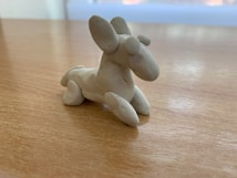 A small, smooth clay sculpture resembling an imaginative creature with large ears, placed on a wooden surface. The sculpture appears detailed with defined features and a simple design.