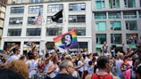 A vibrant parade with people marching and holding rainbow flags. Many participants wear white shirts with a logo. The background includes urban buildings and an American flag.