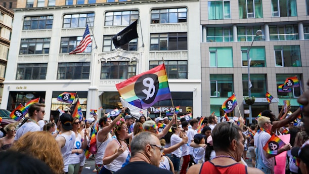 A vibrant parade with people marching and holding rainbow flags. Many participants wear white shirts with a logo. The background includes urban buildings and an American flag.