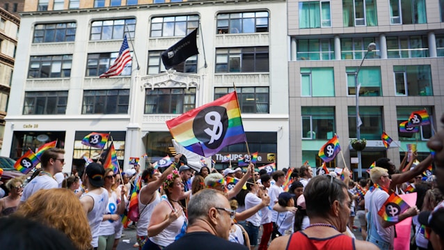 A vibrant parade with people marching and holding rainbow flags. Many participants wear white shirts with a logo. The background includes urban buildings and an American flag.