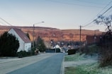 A residential street in a rural area with houses lining both sides. The road is quiet and leads towards rolling hills in the background, illuminated by the soft glow of a setting or rising sun. Telephone poles and wires stretch across the sky, and trees and bushes are seen on either side of the street.