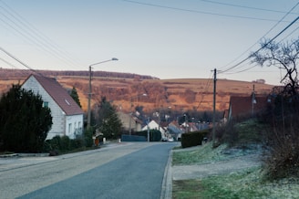 A residential street in a rural area with houses lining both sides. The road is quiet and leads towards rolling hills in the background, illuminated by the soft glow of a setting or rising sun. Telephone poles and wires stretch across the sky, and trees and bushes are seen on either side of the street.