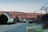 A residential street in a rural area with houses lining both sides. The road is quiet and leads towards rolling hills in the background, illuminated by the soft glow of a setting or rising sun. Telephone poles and wires stretch across the sky, and trees and bushes are seen on either side of the street.